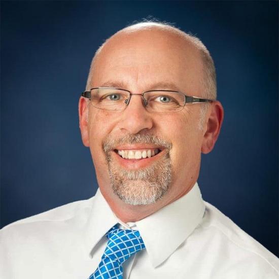 A man with glasses, wearing a white shirt and blue patterned tie, smiles widely in a formal portrait against a blue backdrop.